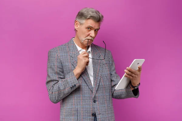 Happy old business man holding white tablet, while staying on violet background.