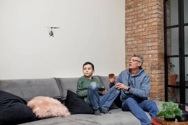 Happy family, old grandfather and cute little boy grandson play  toy plane lay on sofa, funny small grandkid having fun with grandpa fly on airplane laughing at home.