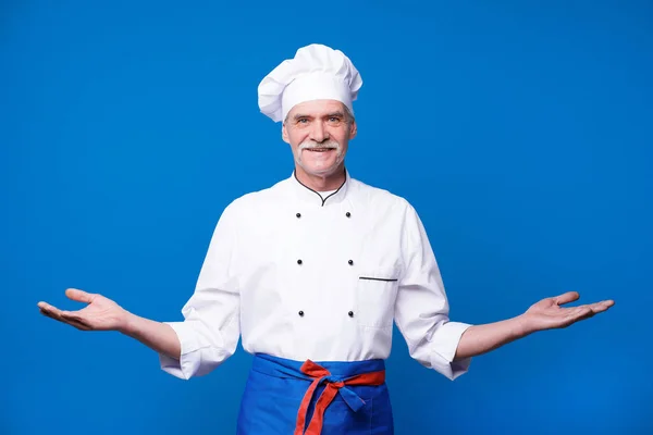 青い背景に孤立した高齢の料理人男はヤシの上にコピースペースを示し 腰に別の手を保持 — ストック写真