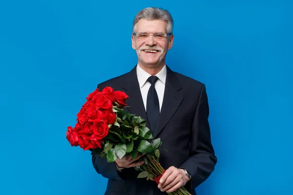Older happy senior man with a bouquet of red rose flowers waiting on a date staying on blue background.
