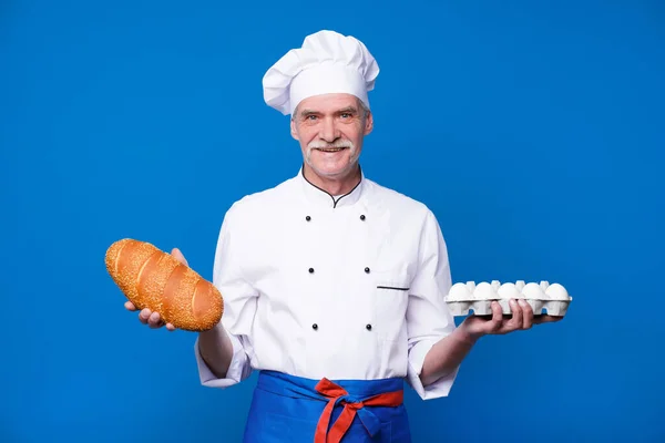 青を背景にポーズをとりながら 卵とパンの入ったバスケットを持つカリスマ的髭シェフの肖像 — ストック写真