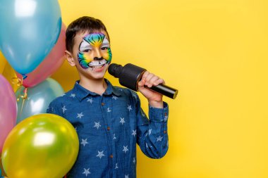 Yüzünde renkli balonlar ve mikrofon olan yakışıklı çocuk sarı arka planda izole bir şekilde duruyor..