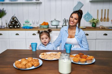 Güzel anne ve küçük kızı mutfakta kahvaltı yapıyorlar. Genç bir aile hafta sonları evde çay içip kruvasan yiyor ve kameraya bakıyor..