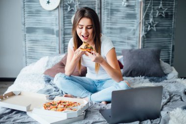 Bir kutu pizzayla yatakta oturan ve pizza yiyen genç bir kızın portresi. Kahvaltıda pizza yiyen mutlu bir kadın..