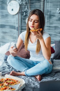 Genç, güzel bir kadın gündelik tarzda giyiniyor pizzayla yatakta oturuyor ve televizyonu açıyor. Otel kahvaltısı!