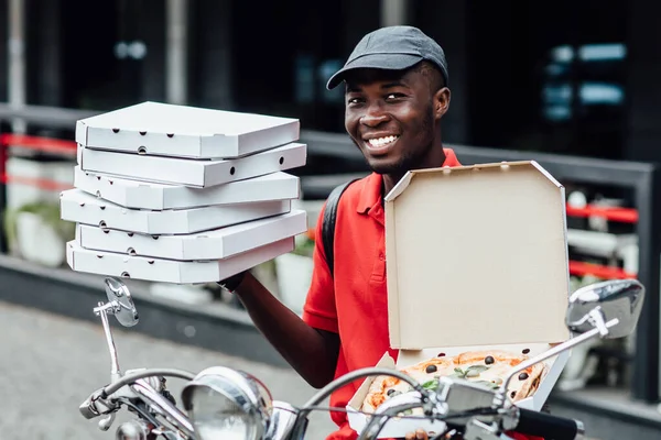 Fast food delivery concept. Happy busy courier or deliveryman shows fresh baked pizza on box, poses on fast scooter, delivers snack. Taste this delicious pizza.