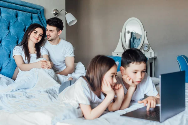 可爱的女孩和男孩躺在床上 带着笔记本电脑看电影 身后是家里的父母 星期一休息一下 — 图库照片