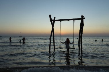 Uzun saçlı kız silueti Phu Quoc adasında deniz batımında salıncakta oturuyor.