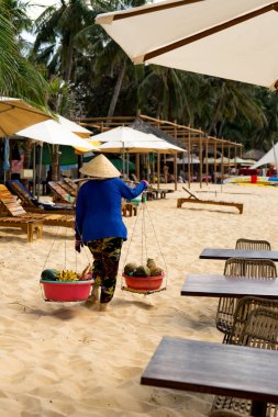 Yaşlı Vietnamlı bir kadın geleneksel Don Ganh hasır meyve sepetiyle sahilde yürüyor. Geleneksel Vietnam şapkası şapkasıyla Phu Quoc adasındaki sokak yemekleri satıcısı..