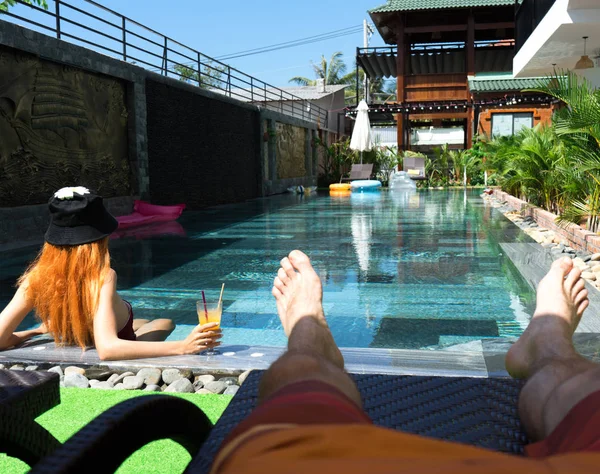 Man chilling near the pool taking selfie of his legs and her girlfiend relaxing in the pool .Couple holidays of young man and woman near the pool of their honeymoon.