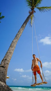 Plaj tatili tropik tarzı palmiye portresi. Plajda sallanan yakışıklı bir adam mavi deniz. Phu Quoc adasında sallanan bir adam, Vietnam. Mutlu adam tropikal palmiye ağacında sallanıyor.