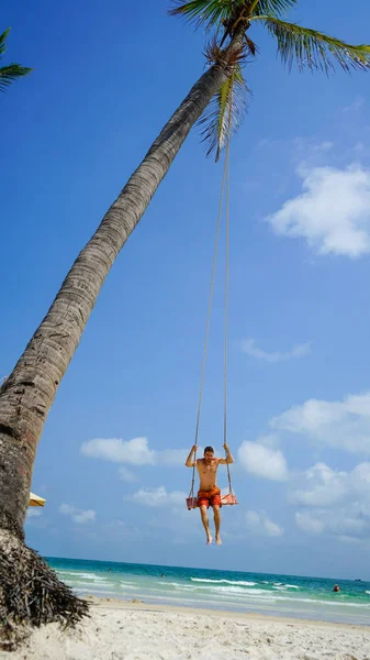 Plaj tatili tropik tarzı palmiye portresi. Plajda sallanan yakışıklı bir adam mavi deniz. Phu Quoc adasında sallanan bir adam, Vietnam. Mutlu adam tropikal palmiye ağacında sallanıyor.