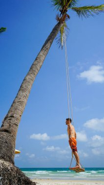 Plaj tatili tropik tarzı palmiye portresi. Plajda sallanan yakışıklı bir adam mavi deniz. Phu Quoc adasında sallanan bir adam, Vietnam. Mutlu adam tropikal palmiye ağacında sallanıyor.