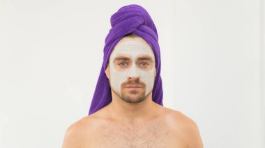 Kafasında mor havlu ve yüzünde kozmetik maske olan komik yakışıklı bir genç adam. Adam yüz kilinden maske takıyor. Kadın Günü, spa ya da sevgililer günü için komik içerikler..