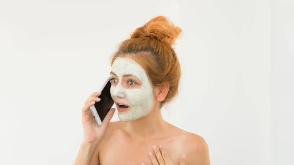 Red haired woman with cosmetic face clay mask in towel after bath talking on the mobile phone
