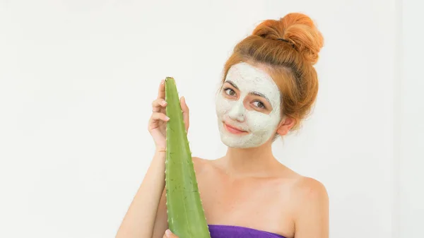 Red haired woman with cosmetic face clay mask on his face wearing  towel after bath and holding alie vera in here hand. Face care with aloe vera.