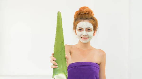 Red haired woman with cosmetic face clay mask on his face wearing  towel after bath and holding alie vera in here hand. Face care with aloe vera.