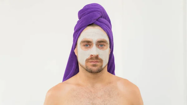 Kafasında mor havlu ve yüzünde kozmetik maske olan komik yakışıklı bir genç adam. Adam yüz kilinden maske takıyor. Kadın Günü, spa ya da sevgililer günü için komik içerikler..