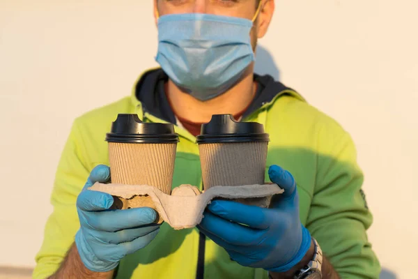 Coffee delivery man in medical rubber gloves and mask holding two cups of coffee. Portrait of young man delivery online or express coffee delivery on quarantine.