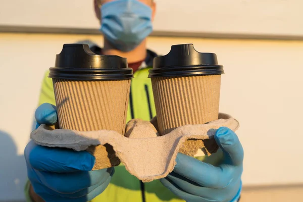 Coffee delivery man in medical rubber gloves and mask holding two cups of coffee. Portrait of young man delivery online or express coffee delivery on quarantine.