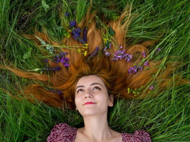 Saçlarında çiçeklerle yeşil yaz çimlerinde uzanan güzel kızıl saçlı kadının yüzünü kapat. Mor elbiseli mutlu kız parkta güneşli çimlerde dinleniyor. Yaz tatili kavramı.