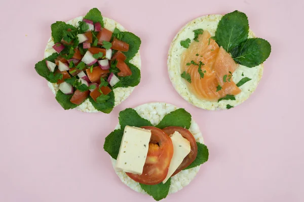 Three rice cakes sandwiches with different healthcare ingredients at the middle on pink background .Rice cakes with tomatoes, salmon, mint and others fit vegetables with copy space in the middle.