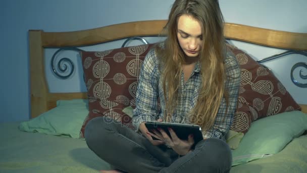 Chica sentada en la cama y sostiene una tableta electrónica — Vídeos de Stock