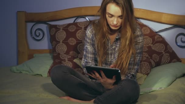 Милая девушка сидит на кровати и смотрит в цифровой планшет — стоковое видео