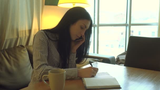 Молодая девушка разговаривает по телефону и что-то пишет в блокноте — стоковое видео