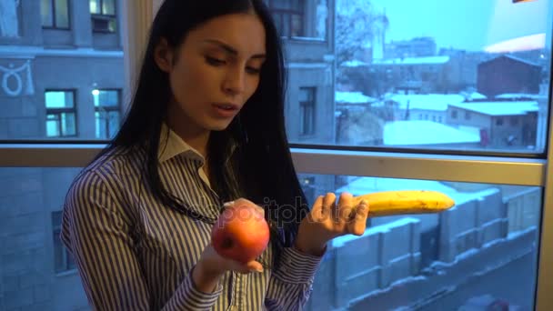 Милая молодая девушка с темными волосами выбирают между яблоком и бананом — стоковое видео