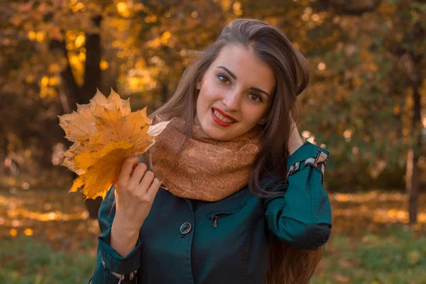 Młoda dziewczyna uśmiechnięta w parku utrzymuje włosy z jednej strony, podczas gdy drugi trzyma liście — Zdjęcie stockowe