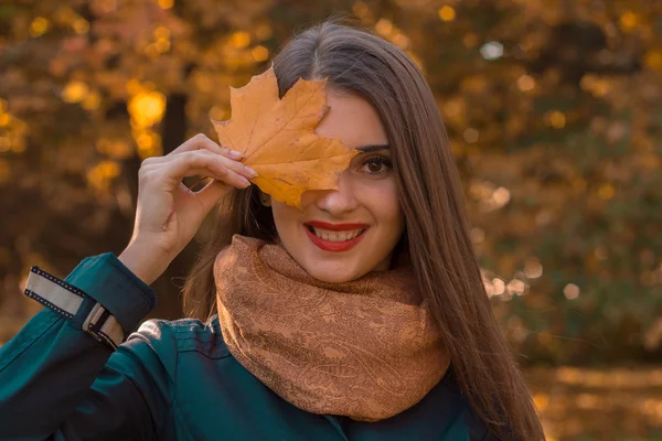 Schönes Mädchen mit roten Lippen im Schal hält ein Ahornblatt vor den Augen und lächelt — Stockfoto