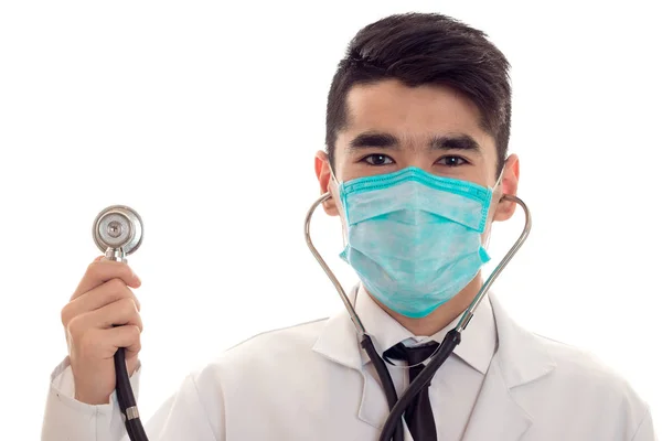 Jeune beau médecin homme posant en uniforme et masque avec stéthoscope isolé sur fond blanc en studio — Photo