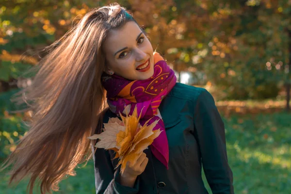 Atractiva joven con el pelo largo en una bufanda rosa mantiene sus hojas en su mano y sonríe — Foto de Stock