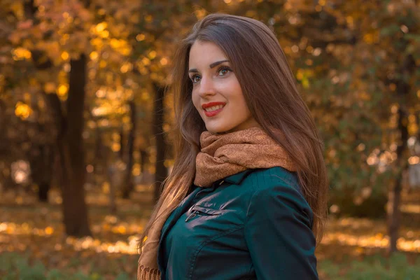 Piękna młoda dziewczyna z szalikiem i ciemne kurtki stoi w parku wygląda od uśmiechów — Zdjęcie stockowe