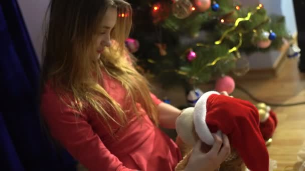 Uma jovem encantadora coloca um chapéu de Papai Noel em seu urso de brinquedo favorito — Vídeo de Stock