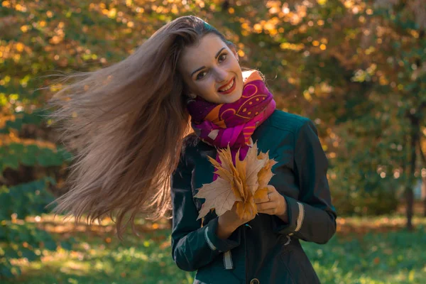 Menina bonita com cabelos longos que voam no vento mantém as folhas em suas mãos e sorri — Fotografia de Stock