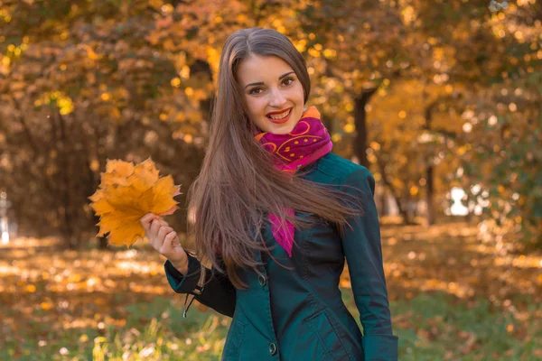 Kul söt flicka i rosa halsduk runt halsen står i parken ser raka och håller bladen i hand — Stockfoto