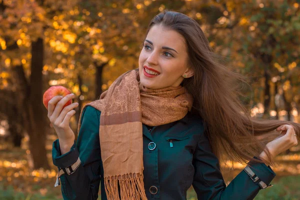 Menina bonita com cabelos longos e um cachecol marrom mantém Apple em sua mão olha para longe sorrisos — Fotografia de Stock