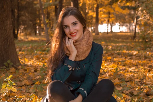 Красивая девушка с красной помадой на губах и теплый шарф сидя в осеннем парке лужайка улыбается — стоковое фото
