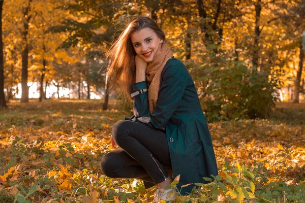 Молода мила дівчина в чорному плащі сидить в парку восени на газоні і посміхається — стокове фото