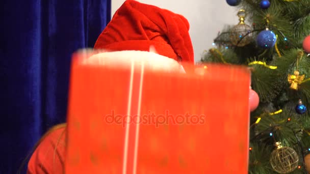 Chica enojada en sombrero de santa se esconde detrás de un regalo de Navidad — Vídeos de Stock