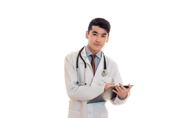 Νεαρά αρσενικά γιατρός με το στηθοσκόπιο με τη στολή και να κάνετε τις σημειώσεις που απομονώνονται σε λευκό φόντο — Φωτογραφία Αρχείου