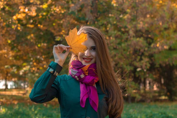 Menina bonita em cachecol mantém folha de outono perto dos olhos e sorrisos — Fotografia de Stock