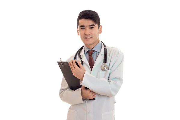 Joven doctor masculino en uniforme con estatoscopio toma notas y sonríe aislado sobre fondo blanco —  Fotos de Stock