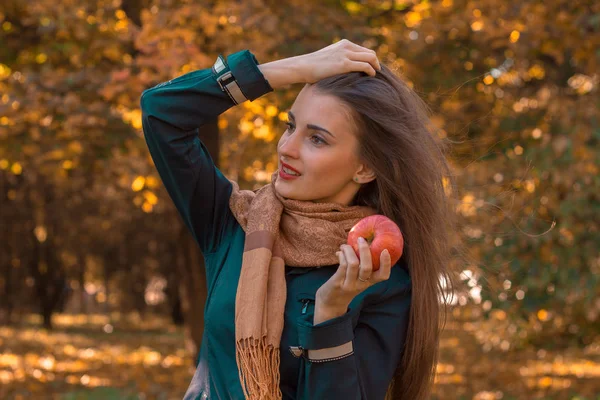 Młoda dziewczyna ubrana w czarną kurtkę i szalik stoi w parku wygląda po stronie trzyma jabłko — Zdjęcie stockowe
