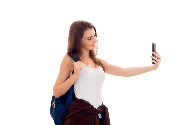 Весёлая юная брюнетка студентка с голубым рюкзаком и мобильным телефоном в руках делает селфи изолированным на белом фоне — стоковое фото