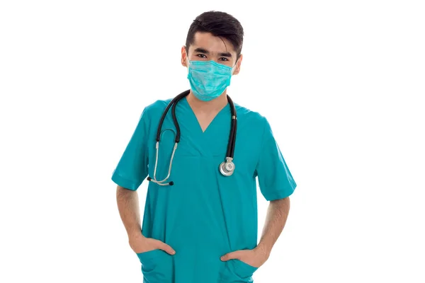 Beau médecin élégant en uniforme bleu avec stéthoscope et masque posant et regardant la caméra isolée sur fond blanc — Photo