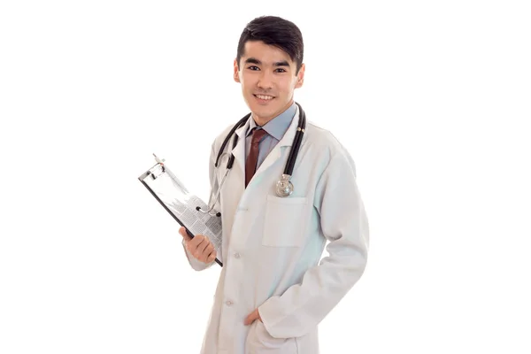 Молода елегантна брюнетка лікар в уніформі зі стетоскопом посміхається на камеру і робить нотатки ізольовані на білому тлі — стокове фото