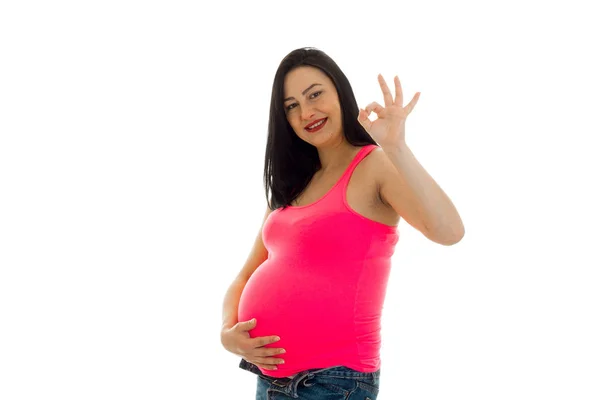 Studio portrait of young pregnant brunette woman in pink shirt touching her belly and showing OK on camera isolated on white background — Stock Photo, Image